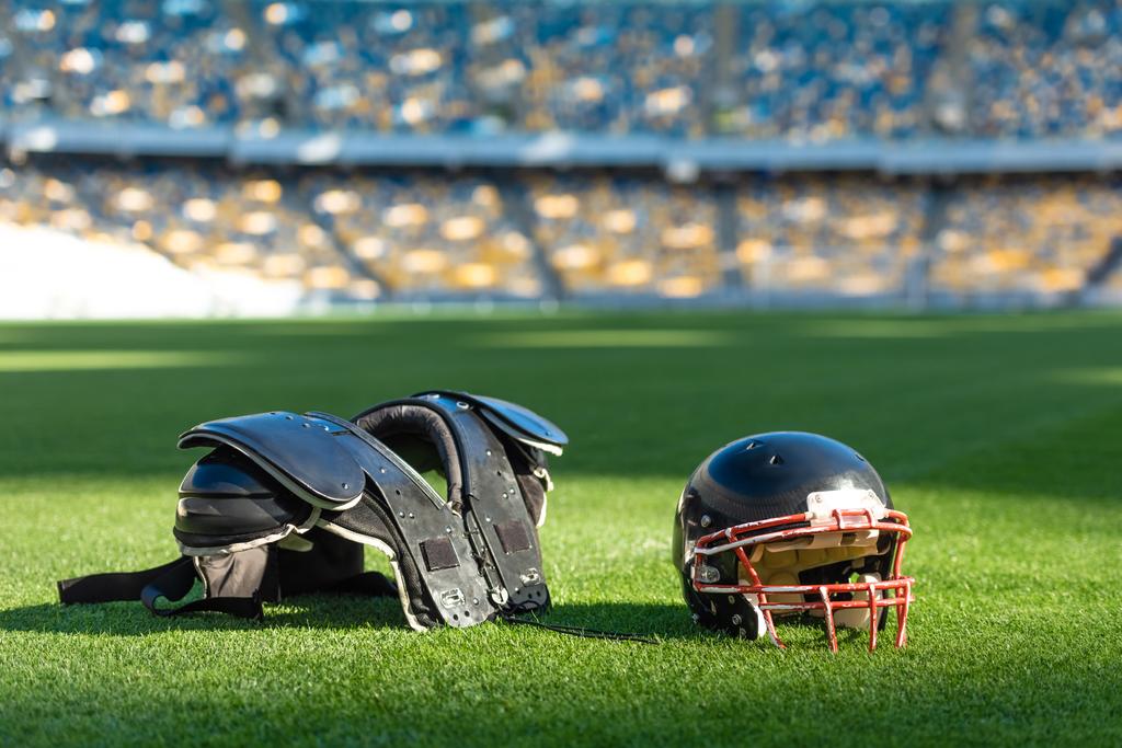 Американский футбольный шлем с защитой груди лежит на зеленой траве стадиона
 - Фото, изображение