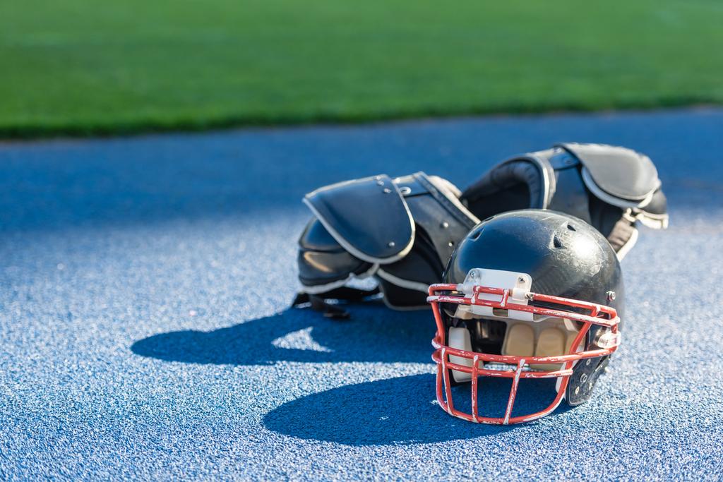 Американский футбольный шлем с защитой груди лежит на синей резиновой поверхности на стадионе
 - Фото, изображение