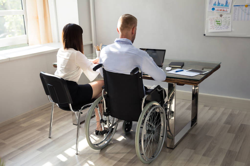 Vista trasera de empresario discapacitado con su compañero de trabajo en el ordenador portátil
 - Foto, Imagen