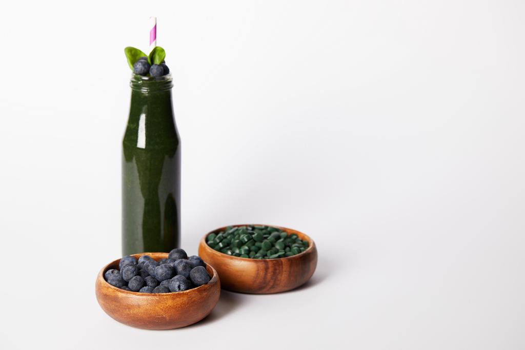 fles van spirulina smoothie met munt verlaat en drinken van stro, kommen met bosbessen en spirulina pillen op grijze achtergrond  - Foto, afbeelding