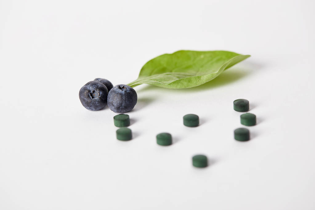 close up view of spirulina pills, blueberries and mint leaf on white background - Photo, Image
