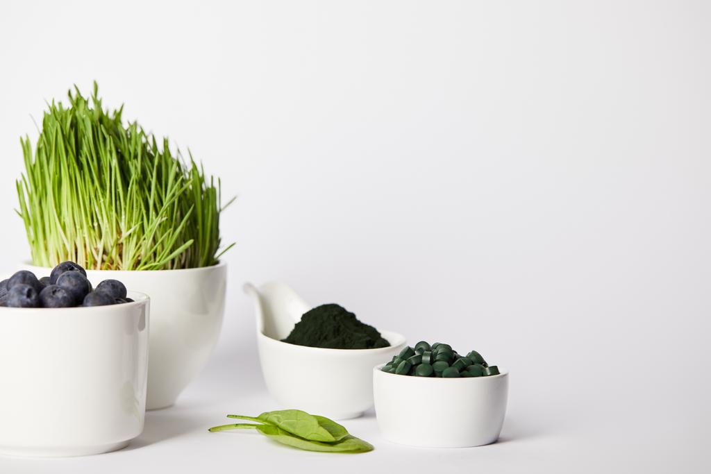 Tassen mit Spirulina-Gras und Blaubeeren, Blätter, Schalen mit Spirulina-Pulver und Spirulina-Pillen auf grauem Hintergrund  - Foto, Bild