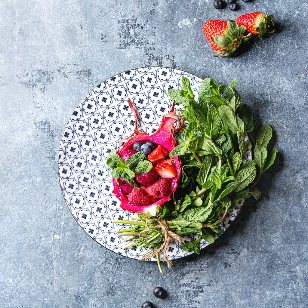 Veganer Obstsalat mit Beeren und Minze in rosa Drachenfrüchten mit Zutaten darüber auf einem Keramikteller vor blauem Hintergrund. Draufsicht, Raum. Gesunde Ernährung - Foto, Bild