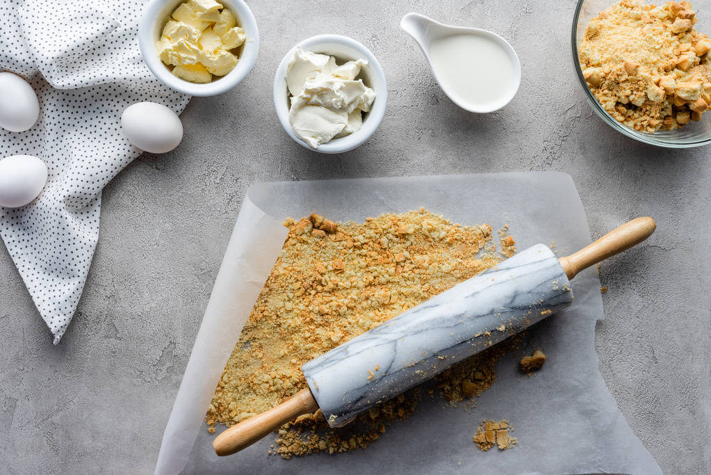 la puesta plana con rodillo, galletas trituradas, huevos de pollo crudos y mantequilla para hacer pastel en la superficie gris
 - Foto, imagen