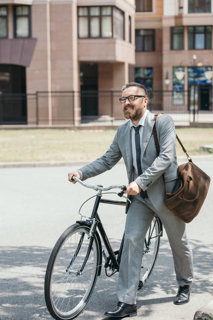 handsome businessman in grey suit and glasses walking with bike on street in city - Photo, Image