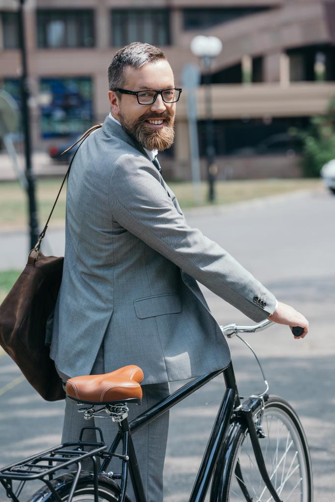 sorridente bonito empresário de pé com bicicleta e olhando para a câmera na rua na cidade
 - Foto, Imagem