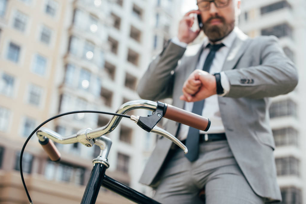 επιλεκτική εστίαση του επιχειρηματία μιλώντας στο smartphone και να βλέπουν το Ρολόι χεριού ενώ κάθεται στο ποδήλατο  - Φωτογραφία, εικόνα