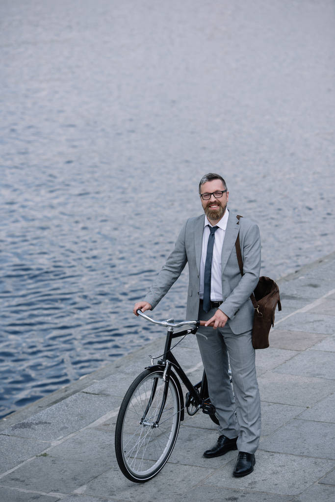 красивый бизнесмен с кожаной сумкой, стоящий на набережной с велосипедом
 - Фото, изображение