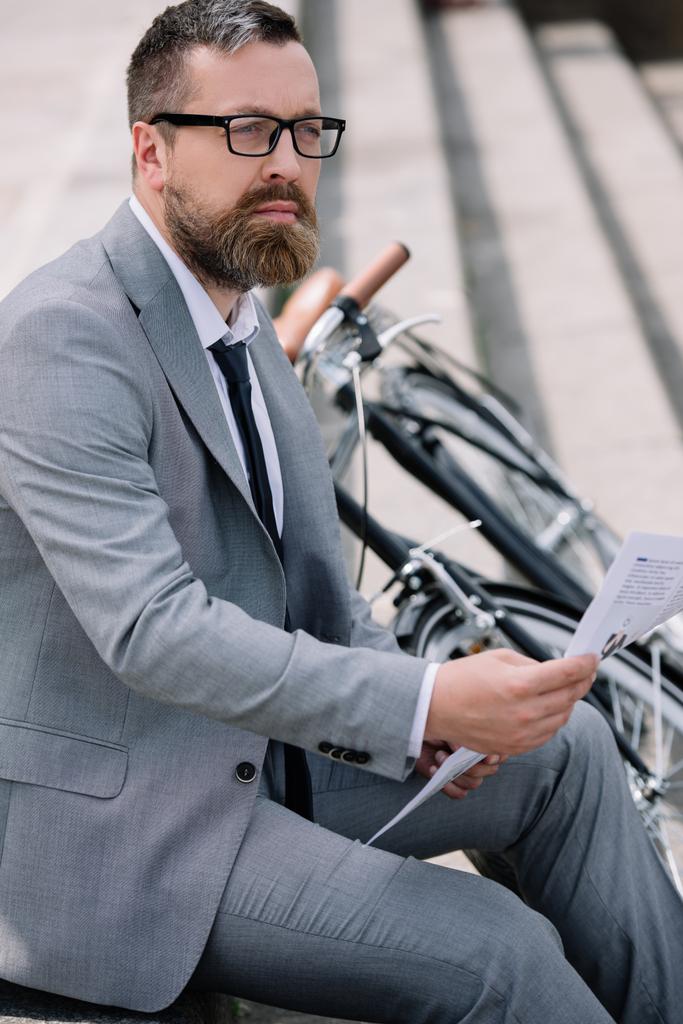 mietteliäs parrakas liikemies lukemassa sanomalehteä portaissa polkupyörällä
 - Valokuva, kuva
