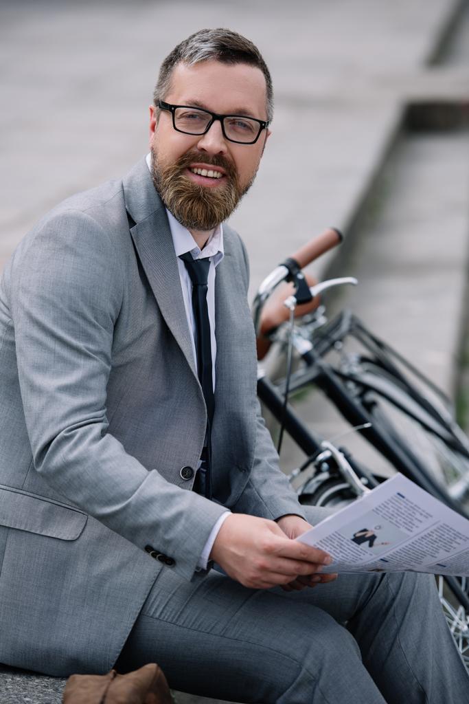 schöner bärtiger Geschäftsmann mit Zeitung sitzt mit Fahrrad auf der Treppe - Foto, Bild