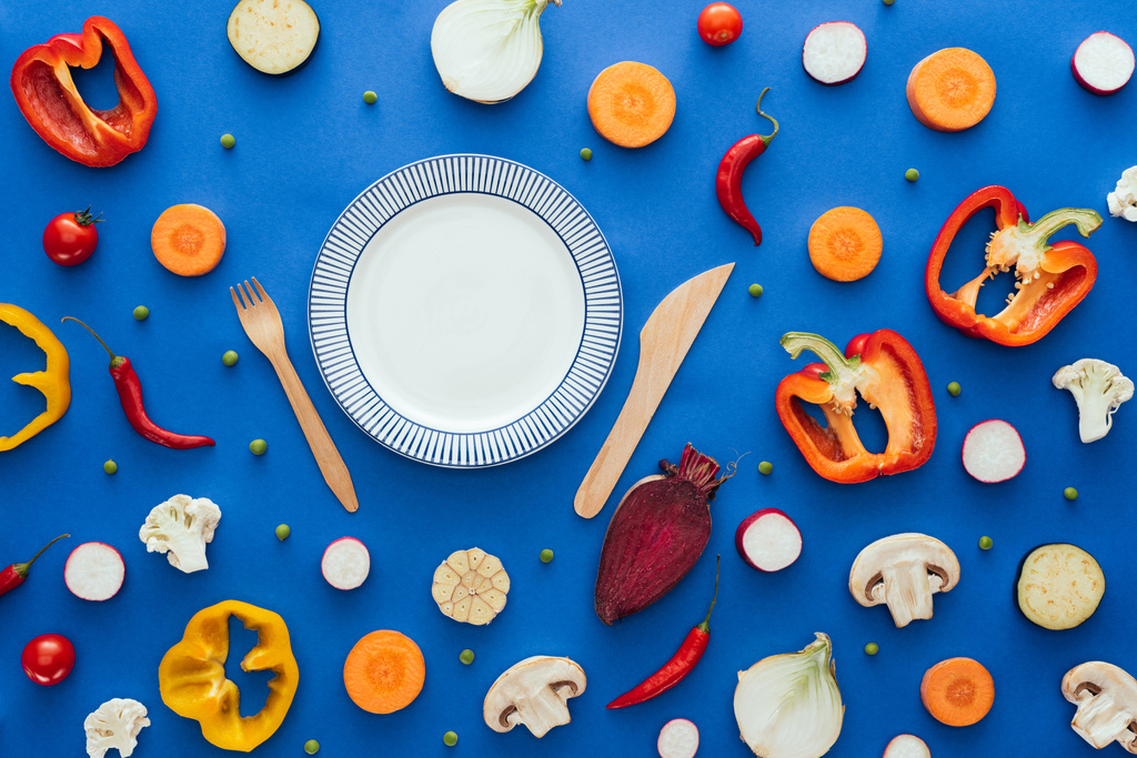 вид сверху на пустую тарелку с вилкой и ножом и различные свежие нарезанные овощи на голубом
 - Фото, изображение