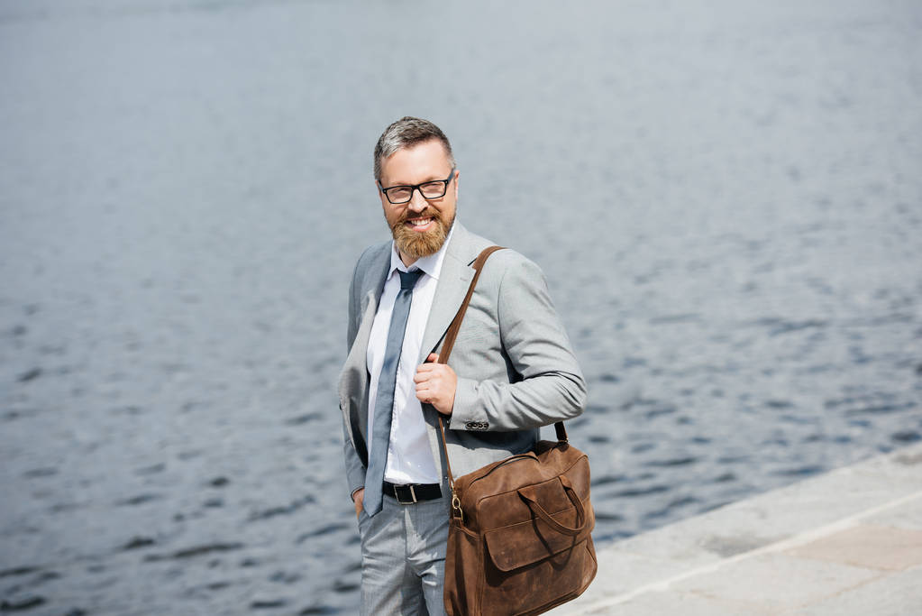 hombre de negocios guapo con bolsa de cuero caminando en muelle cerca del río
  - Foto, imagen