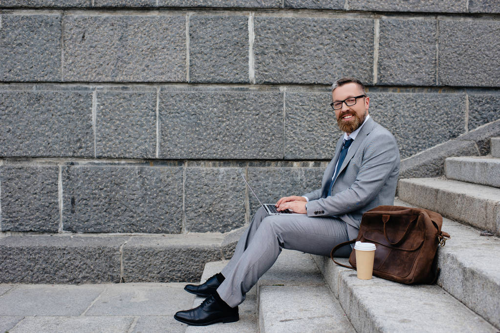 homem de negócios com saco e xícara descartável de café usando laptop e sentado em escadas
 - Foto, Imagem