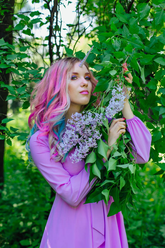 Fata ragazza unicorno con i capelli arcobaleno e trucco lucido tenendo fiori lilla
. - Foto, immagini