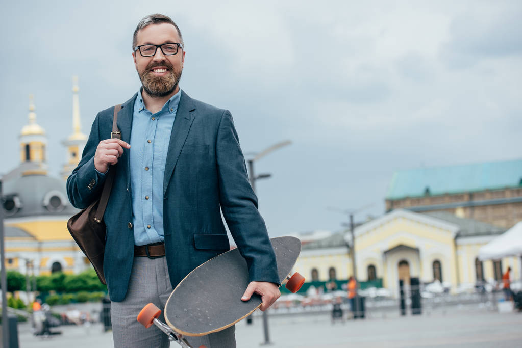 smiling bearded man with leather bag and longboard walking in city - Photo, Image
