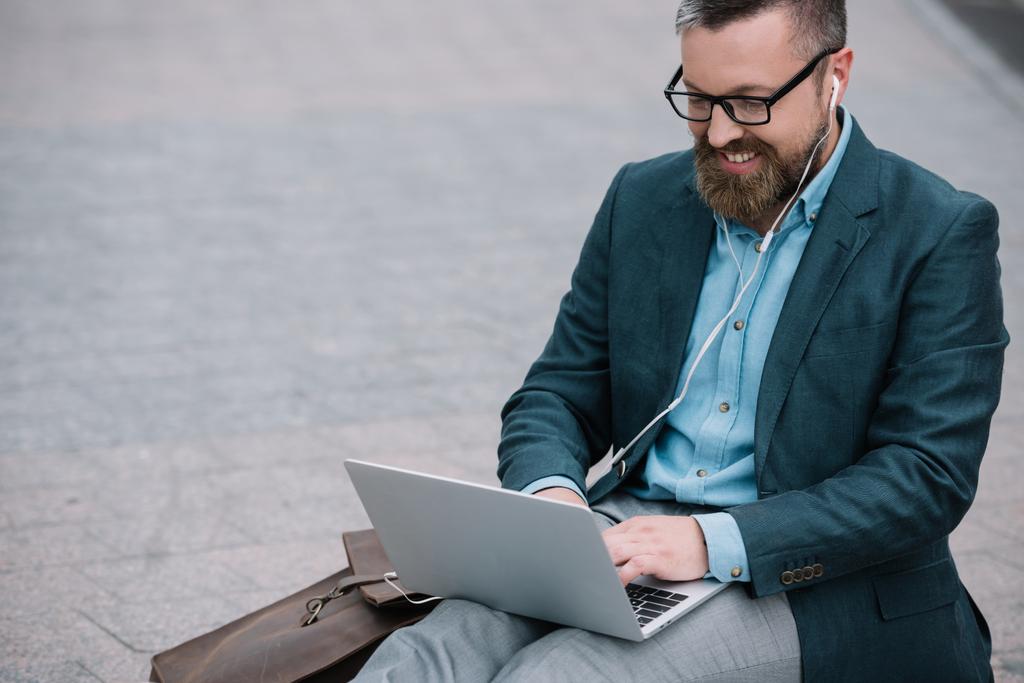 стильний бородатий чоловік використовує ноутбук з навушниками та сидить у місті
 - Фото, зображення
