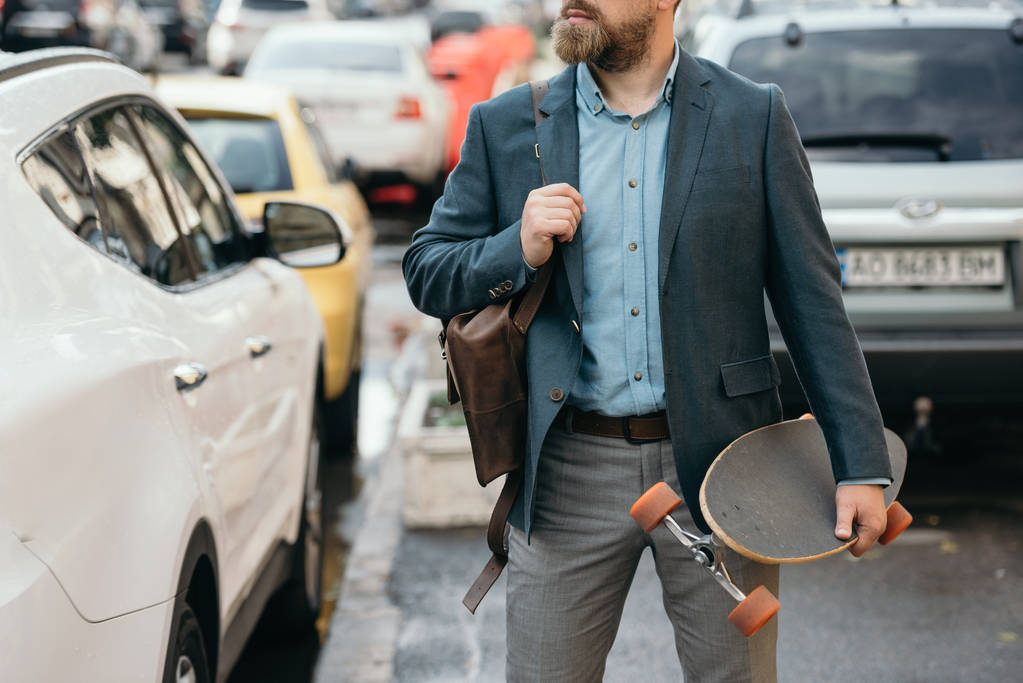 oříznutý pohled na muže s koženou tašku a longboard v městě s auty - Fotografie, Obrázek