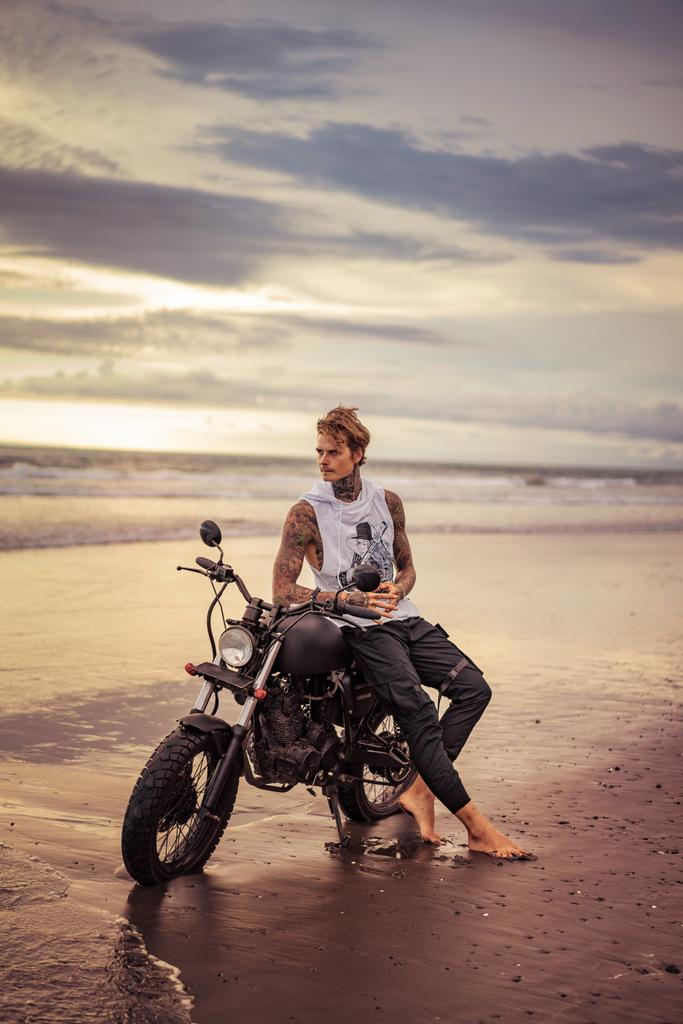 hombre tatuado guapo sentado en moto en la playa del océano y mirando hacia otro lado
 - Foto, Imagen