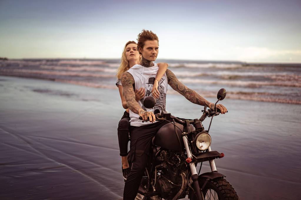joven elegante pareja tatuada a caballo motocicleta en la playa del océano
  - Foto, Imagen