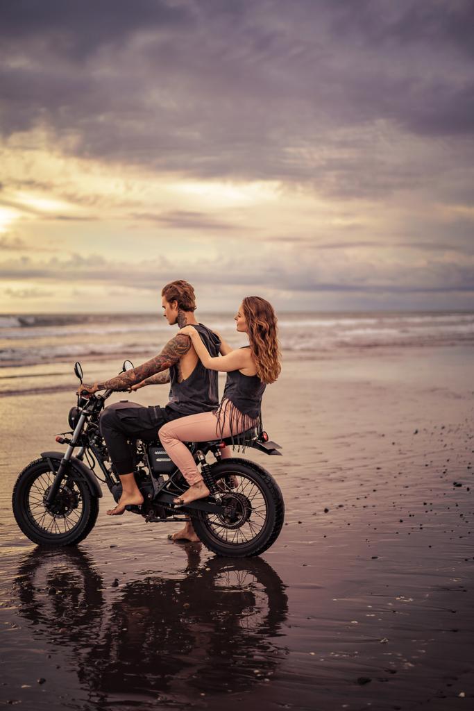Widok z boku para siedzi na motocyklu na plaży podczas sunrise  - Zdjęcie, obraz
