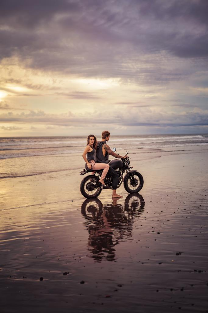 Freund und Freundin sitzen bei Sonnenaufgang und bewölktem Wetter auf dem Motorrad am Strand - Foto, Bild