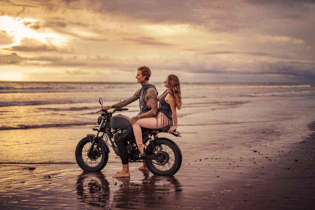 novio y novia tatuados sentados en motocicleta en la playa durante el amanecer
  - Foto, imagen