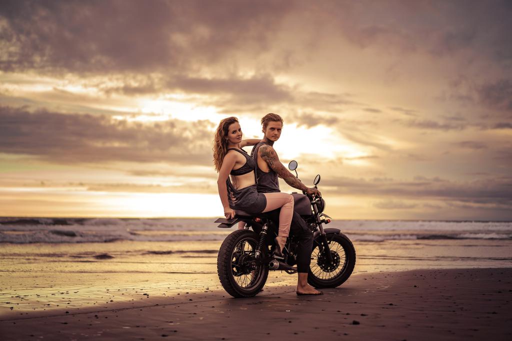 vriend en vriendin zittend op motorfiets op strand tijdens zonsopgang en de camera kijken - Foto, afbeelding