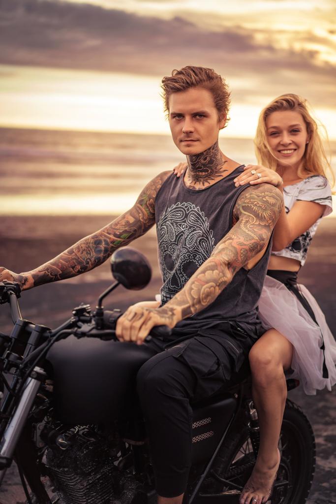 νέοι ετεροφυλόφιλο ζευγάρι κάθεται σε μοτοσικλέτα και να βλέπουν τα φωτογραφικών μηχανών στην παραλία - Φωτογραφία, εικόνα