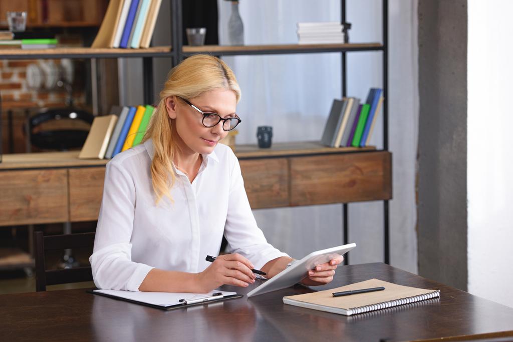 зосереджена жінка-консультант в окулярах, дивлячись на екран цифрових планшетів за столом в офісі
  - Фото, зображення