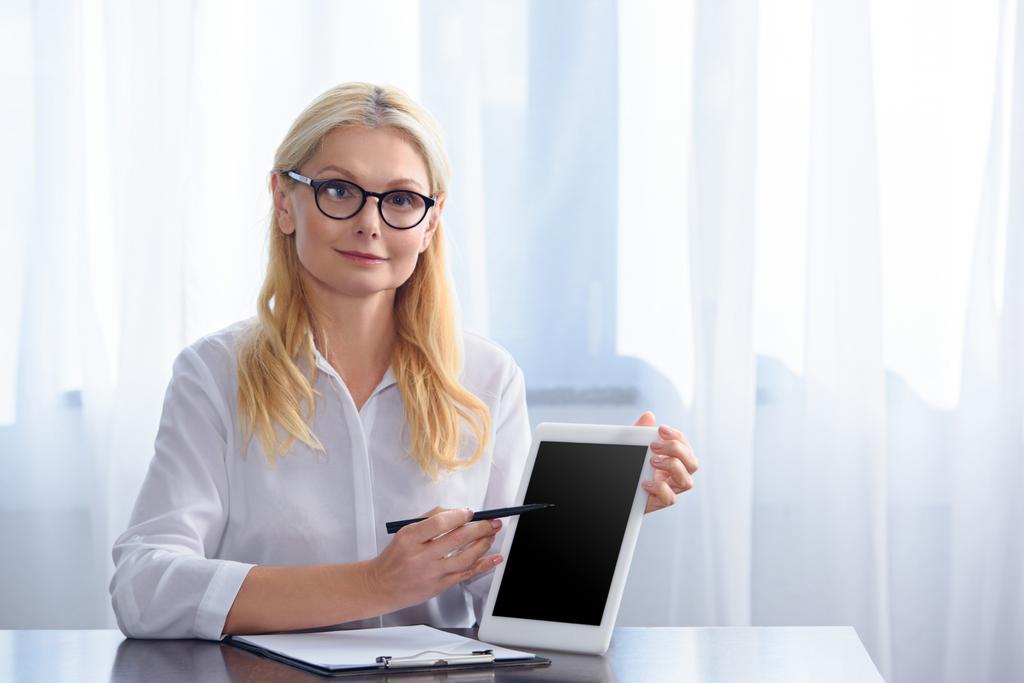 glimlachend vrouwelijke raadgever in brillen wijzen op digitale tablet met leeg scherm aan tafel in kantoor  - Foto, afbeelding