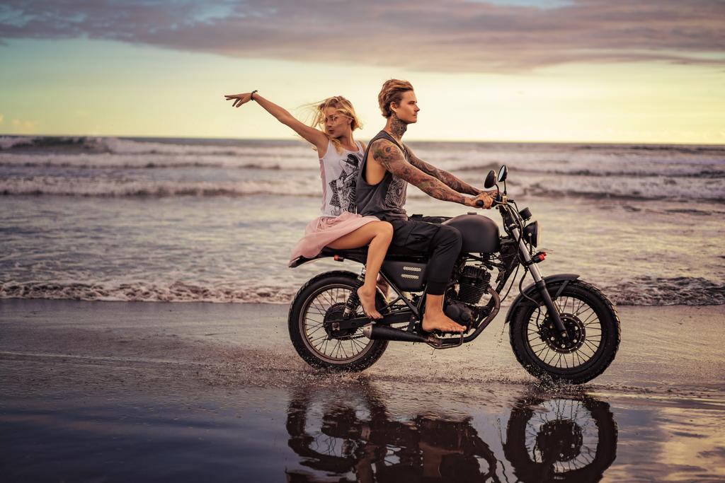 happy boyfriend and girlfriend riding motorbike on seashore during sunrise - Photo, Image