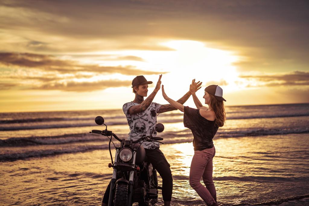 pár dávat vysoké pět poblíž motocykl na pláži u oceánu během východu slunce - Fotografie, Obrázek