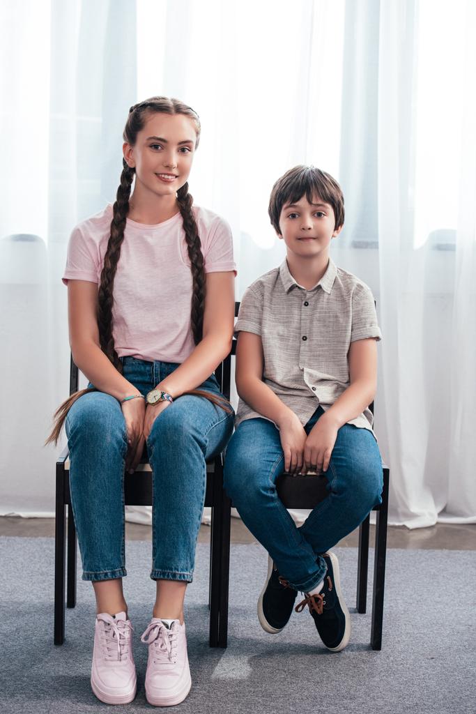 lachende tienermeisje met broer zitten op stoelen en camera thuis te kijken  - Foto, afbeelding