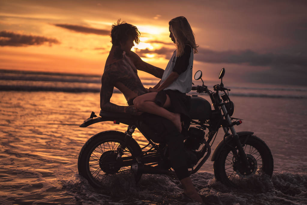 vriendin zit op shirtless vriendje op motor op strand tijdens zonsondergang - Foto, afbeelding