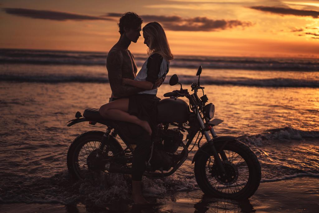 verleidelijke paar knuffelen op motor op strand tijdens zonsondergang - Foto, afbeelding