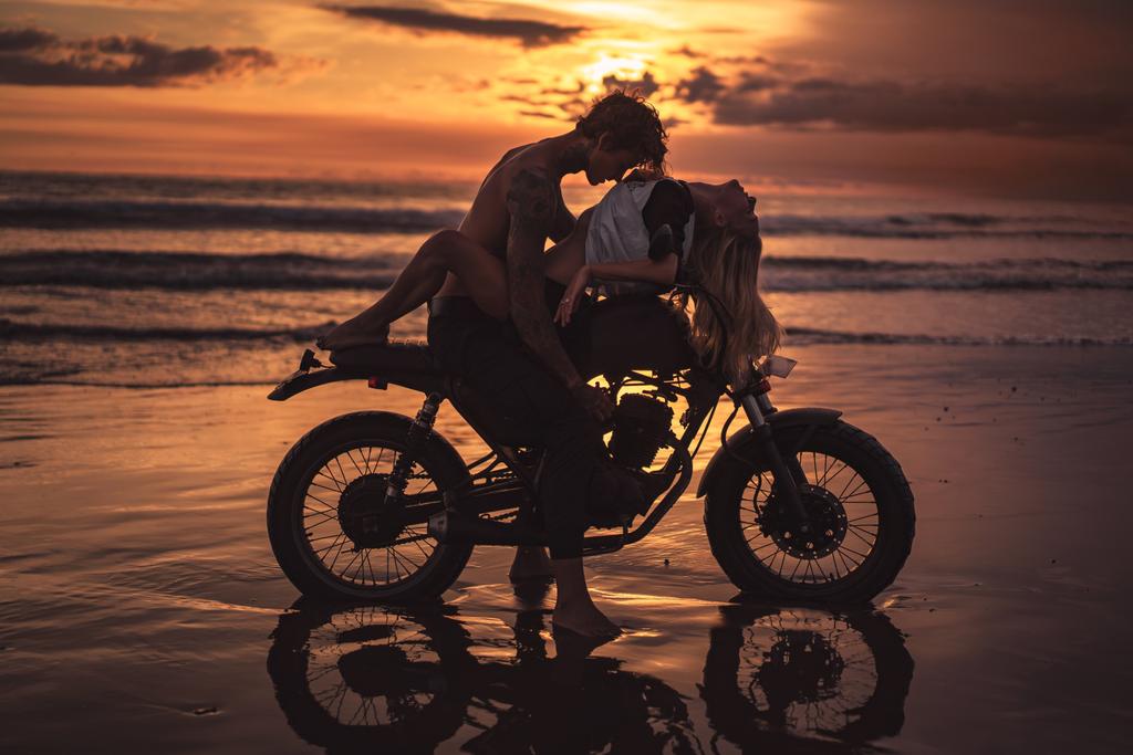Σέξι ζευγάρι αγκαλιάζει τη μοτοσικλέτα στην παραλία κατά τη διάρκεια του ηλιοβασιλέματος - Φωτογραφία, εικόνα