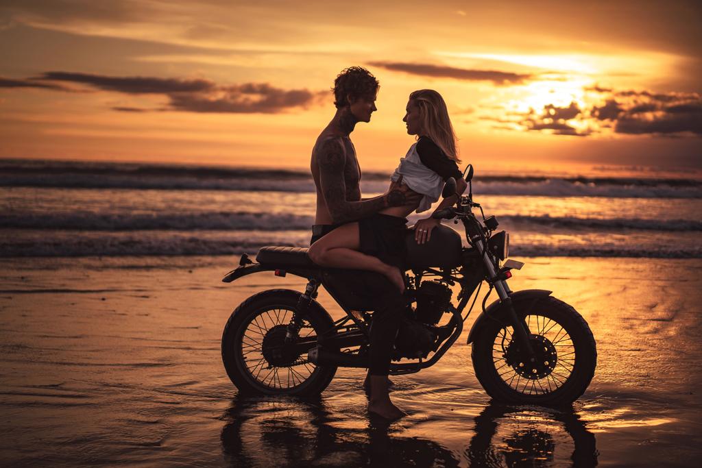 Sinnliches Paar, das sich bei Sonnenuntergang auf dem Motorrad am Strand umarmt - Foto, Bild