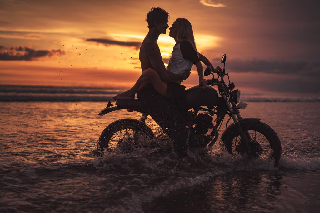 Παθιασμένος ζευγάρι αγκαλιάζει τη μοτοσικλέτα στην παραλία κατά τη διάρκεια του ηλιοβασιλέματος - Φωτογραφία, εικόνα