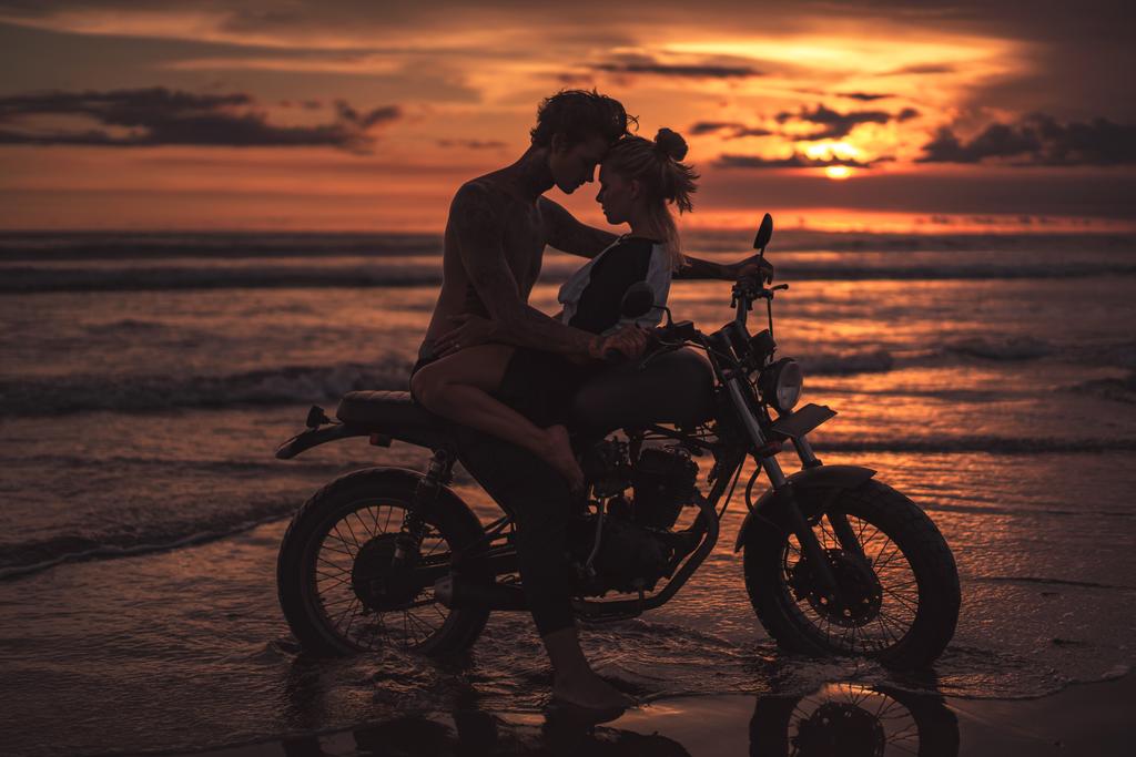страстная пара, обнимающая и трогающая лбы на мотоцикле на пляже во время заката
  - Фото, изображение