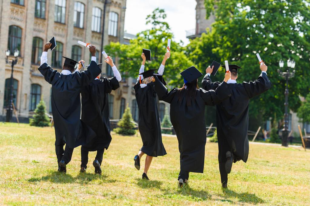 Blick von hinten auf feiernde junge Absolventen in Umhängen, die durch den Universitätsgarten spazieren - Foto, Bild