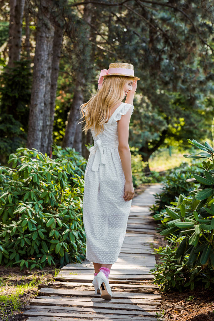 zadní pohled na krásnou dívku v bílé šaty a proutěný klobouk na dřevěný chodník v parku - Fotografie, Obrázek