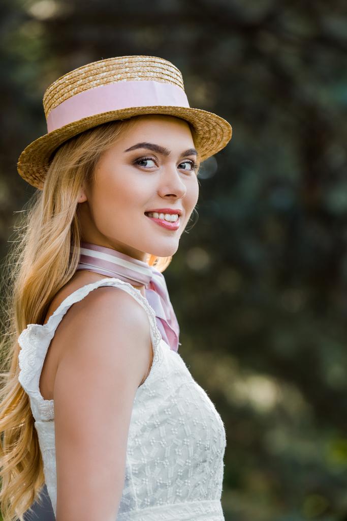 vista lateral de la hermosa mujer joven en sombrero de mimbre sonriendo a la cámara en el parque
 - Foto, imagen