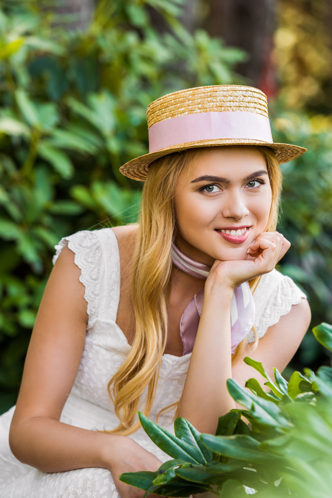 portrét krásné mladé blond ženy v proutěné klobouk s mašlí se usmívá na kameru mezi zelenými rostlinami - Fotografie, Obrázek