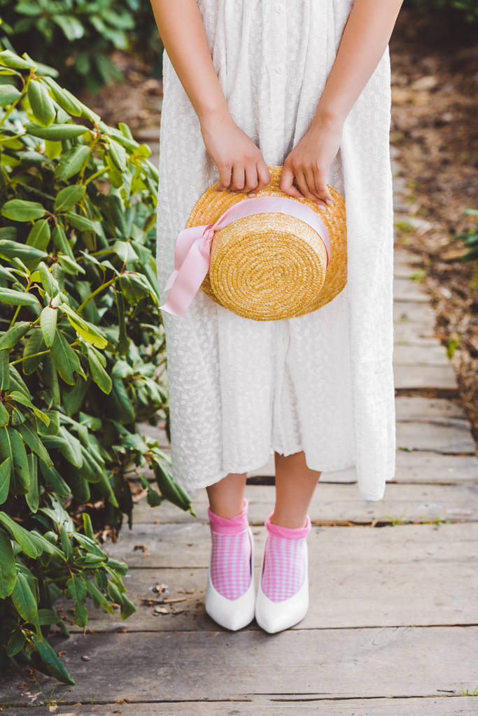 リボン付き籐帽子を保持している白いドレスの少女のクロップ撮影 - 写真・画像