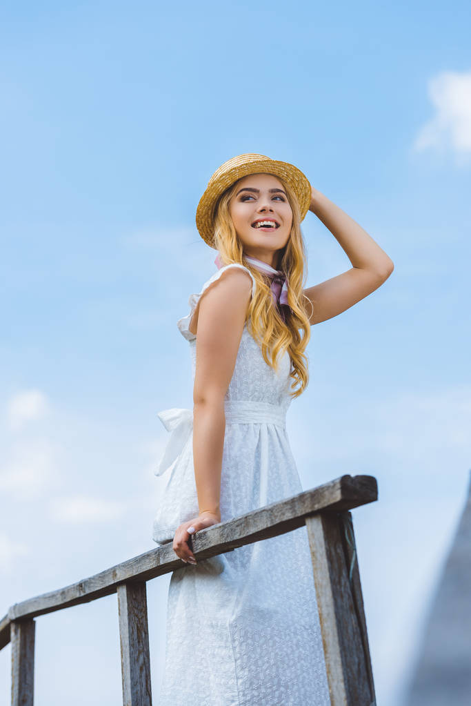 vue à angle bas de fille blonde heureuse en osier chapeau souriant et regardant loin contre le ciel bleu
  - Photo, image