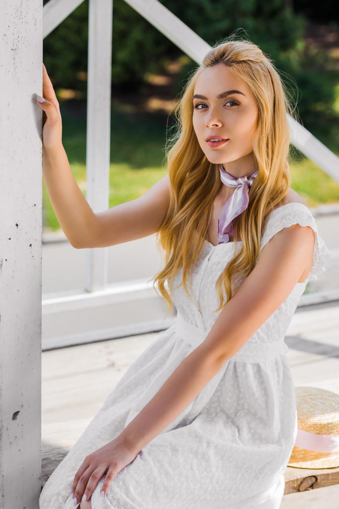 schöne junge blonde Frau im weißen Kleid sitzt auf Bank und lächelt in die Kamera - Foto, Bild