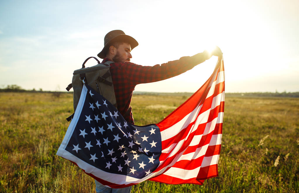4 июля. Американский флаг. Патриотический праздник. Путешественник с флагом Америки. На человеке шляпа, рюкзак, рубашка и джинсы. Прекрасный солнечный свет. Американский стиль
.  - Фото, изображение