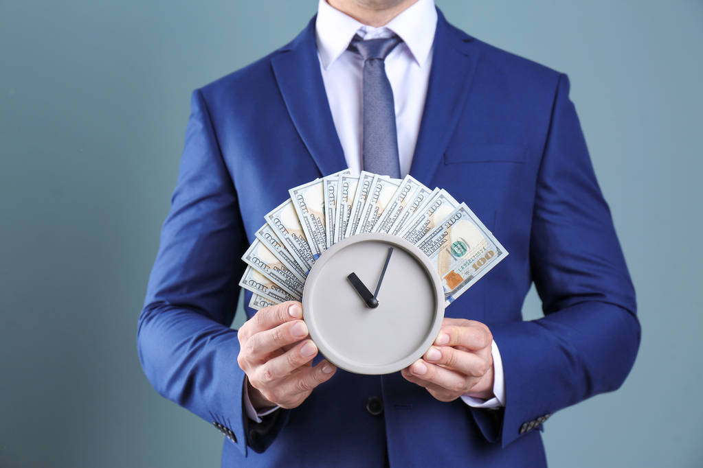 Young businessman holding clock and money on color background. Time management - Photo, Image