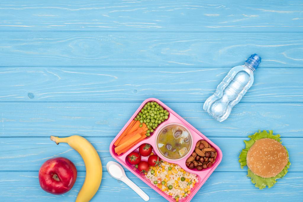 Draufsicht auf Tablett mit Mittagessen für Schulkinder, Flasche Wasser und Obst auf blauer Holzoberfläche - Foto, Bild