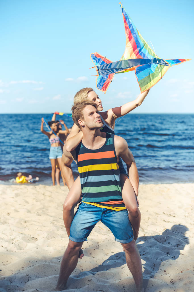 selective focus of couple with colorful kite  with multiracial friends behind spending time on sandy beach - Photo, Image